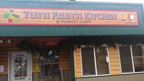 Sign for "Tutti Fruitti Kitchen & Market Farm" on a wooden building, featuring produce and restaurant elements.