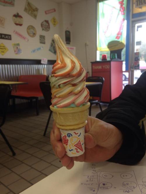 A hand holding a colorful swirl ice cream cone with orange, yellow, and brown stripes in a casual ice cream shop.