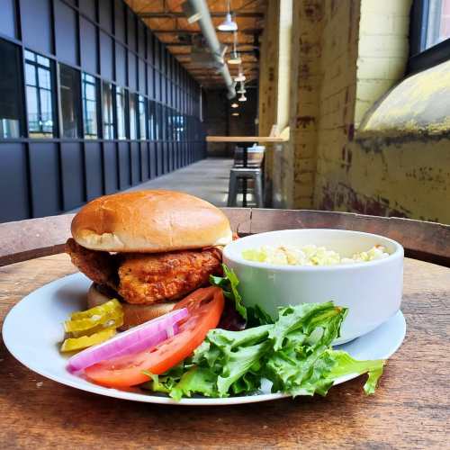 A crispy chicken sandwich with lettuce, tomato, pickles, and a side of coleslaw on a wooden table in a spacious setting.