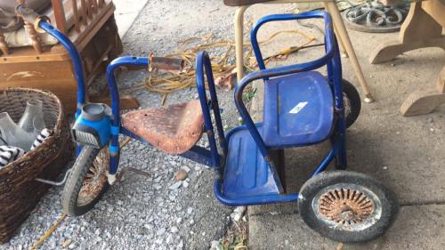A rusty blue tricycle with a missing seat, leaning against a wall on a gravel surface, surrounded by various items.