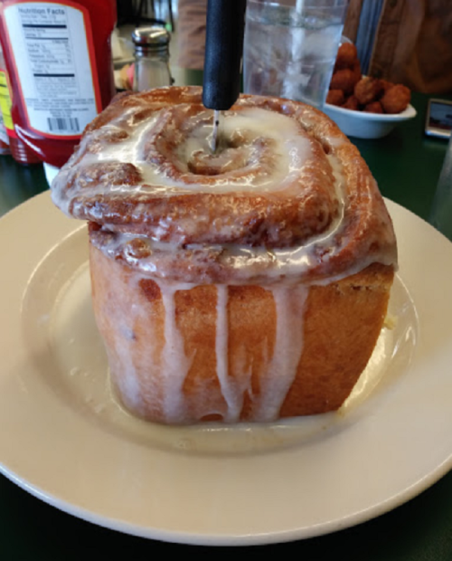 A large, gooey cinnamon roll with icing, served on a plate with a knife inserted into it.