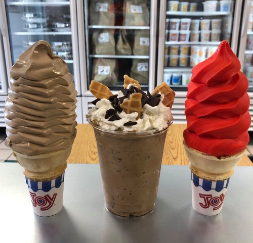 Two soft serve ice cream cones, one chocolate and one red, flank a cup of chocolate milkshake topped with whipped cream and toppings.