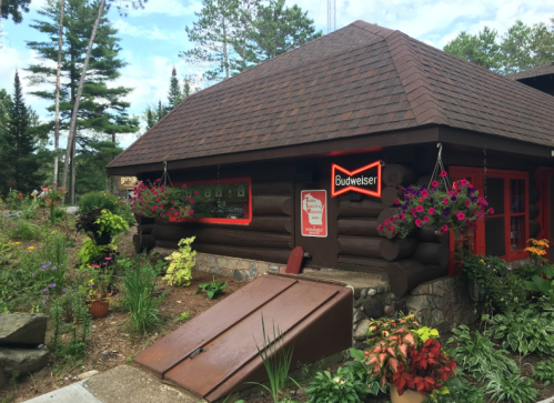 A rustic log cabin with a Budweiser sign, surrounded by colorful flowers and greenery under a blue sky.