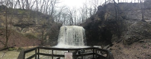 A waterfall cascades over rocky cliffs, surrounded by bare trees and a wooden viewing platform.