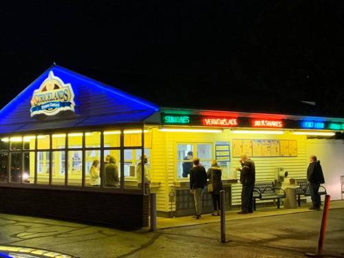 A brightly lit ice cream shop at night, with colorful signs and customers waiting outside.