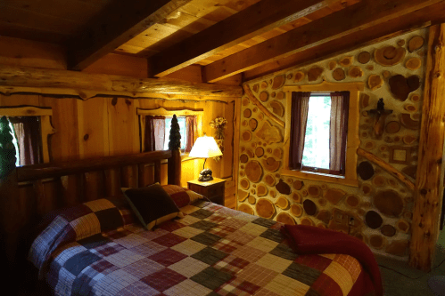 Cozy cabin bedroom with a wooden bed, plaid bedding, and log walls, featuring a lamp and window with curtains.