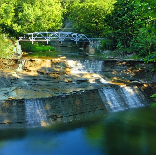 A serene waterfall cascades over rocky ledges, with a bridge crossing a calm river surrounded by lush greenery.