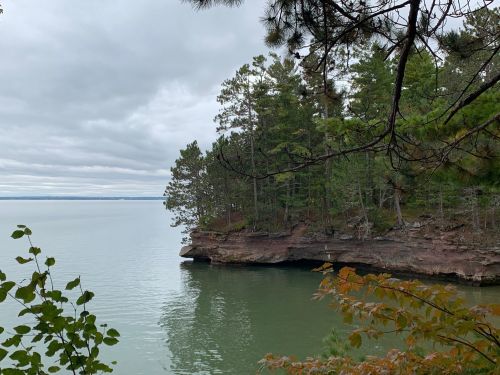 A serene view of a calm lake surrounded by lush green trees and rocky cliffs under a cloudy sky.