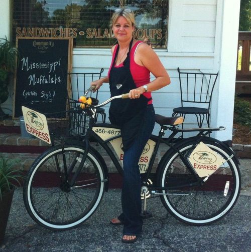 A woman in an apron stands next to a bicycle with signs for a sandwich shop, smiling in front of a restaurant.