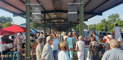 A bustling outdoor market with vendors, tents, and a crowd of people shopping and socializing under a covered area.