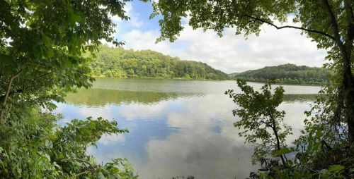 A serene lake surrounded by lush greenery and trees, reflecting the sky and clouds above.