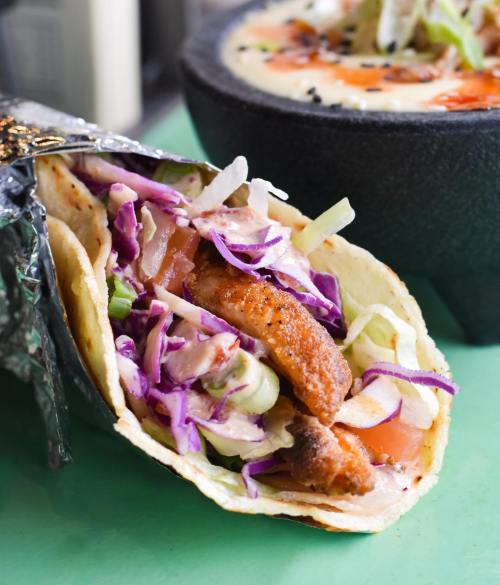 A close-up of a taco filled with grilled meat, cabbage, and tomatoes, with a bowl of soup in the background.