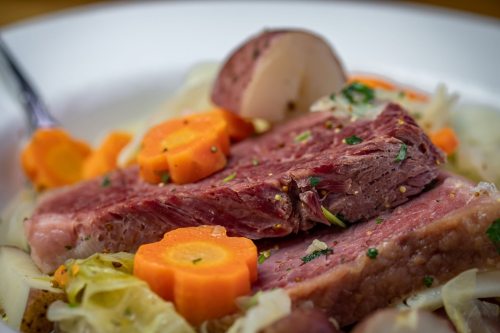 A close-up of a plate featuring sliced corned beef, carrots, potatoes, and cabbage, garnished with herbs.