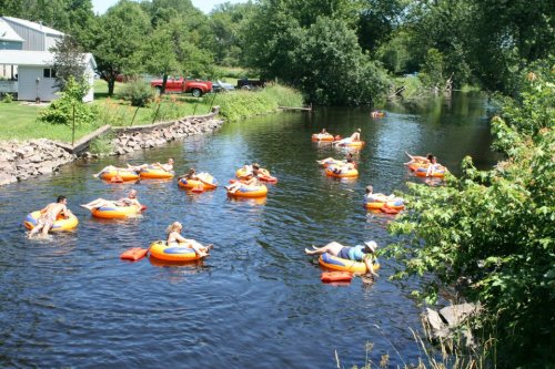 People relaxing on orange inner tubes in a calm river, surrounded by greenery and a few houses in the background.