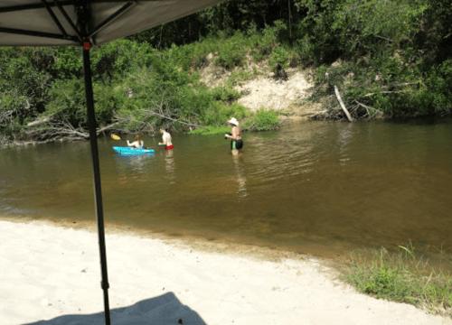 Two people wade in a shallow river while another sits in a kayak. A sandy beach and trees are visible in the background.