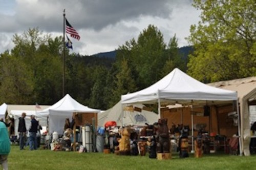 Tents at an outdoor market with various crafts and goods, set against a backdrop of trees and a cloudy sky.