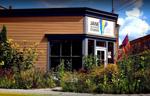 A colorful art studio with a sign reading "Jane Wilkinson Studios," surrounded by vibrant flowers and greenery.