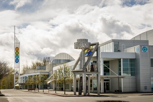 Modern building with large glass windows, colorful signage, and a sculpture in front, under a cloudy sky.