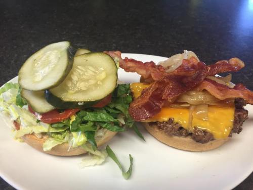 A plate with a burger topped with cheddar cheese, bacon, and onions, alongside a fresh bun with lettuce, tomato, and pickles.