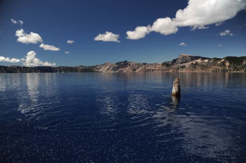 A serene lake with calm blue water, surrounded by cliffs and clouds, featuring a lone wooden stump in the foreground.