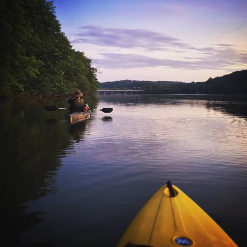 A person kayaking on a calm river at sunset, with lush greenery along the banks and a bridge in the distance.