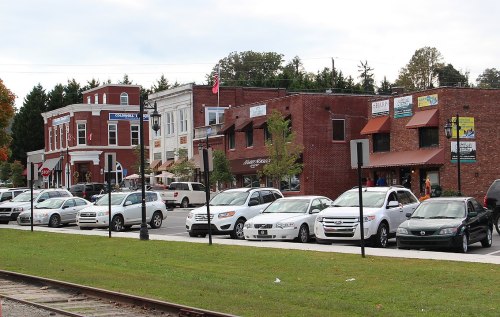 A row of brick and storefront buildings with parked cars along a street, surrounded by trees and a cloudy sky.