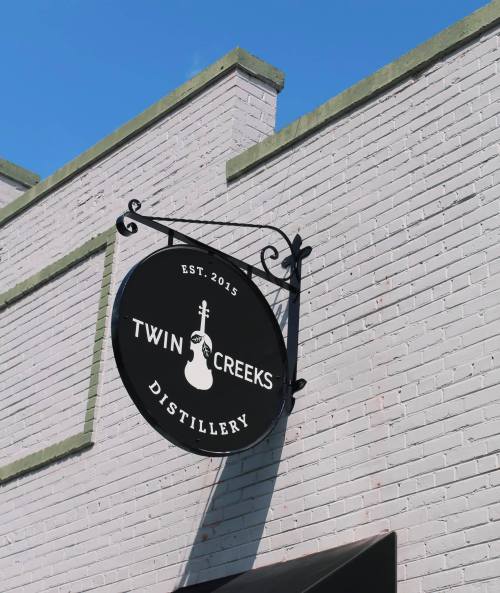 Sign for Twin Creeks Distillery, established in 2015, featuring a guitar graphic against a white brick wall.