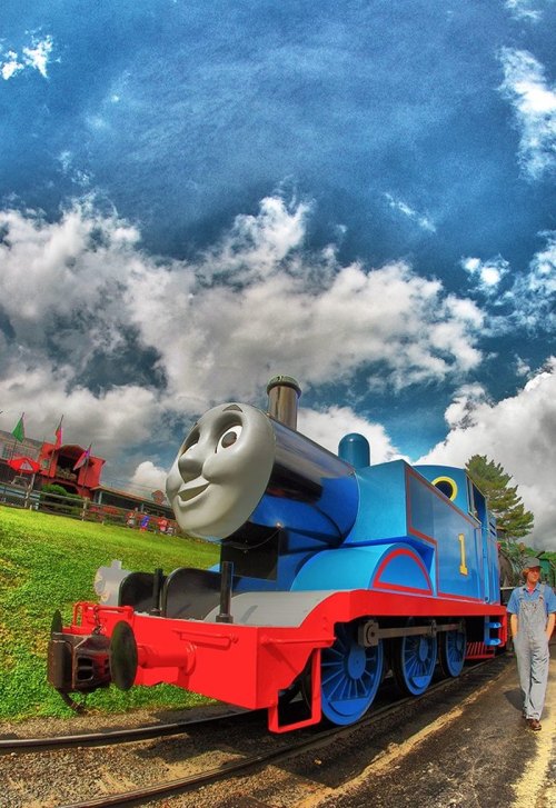 A blue steam engine with a smiling face, parked on tracks under a cloudy sky, with a person in overalls nearby.