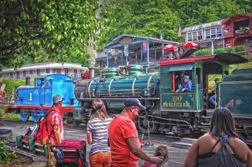 A colorful train station scene with a blue cartoon train and a vintage steam locomotive, surrounded by people and greenery.