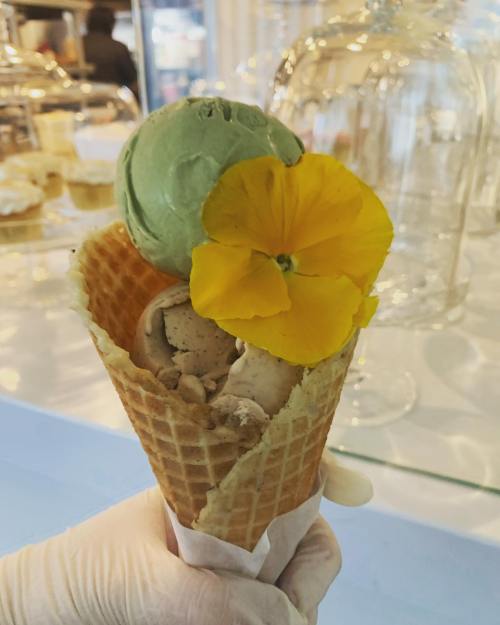 A hand holding a waffle cone filled with two ice cream scoops and a yellow flower on top.