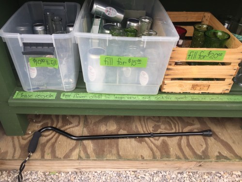 Baskets and bins filled with glassware for sale, with a long tool resting on the ground. Recycling message visible.