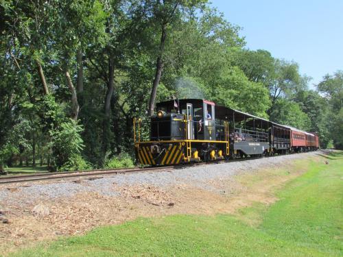 A black and yellow locomotive pulls a passenger car along a tree-lined track on a sunny day.