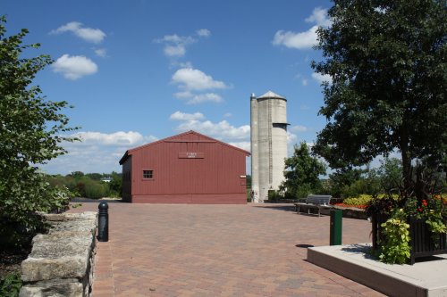 A red barn and a silo stand against a blue sky with fluffy clouds, surrounded by greenery and a paved walkway.