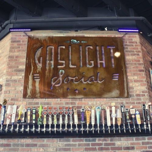 Sign for "The Gaslight Social" above a wall of various beer taps in a rustic bar setting.