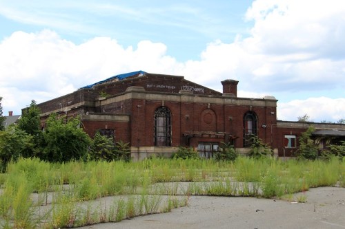 Abandoned brick building surrounded by overgrown grass and weeds under a cloudy sky.
