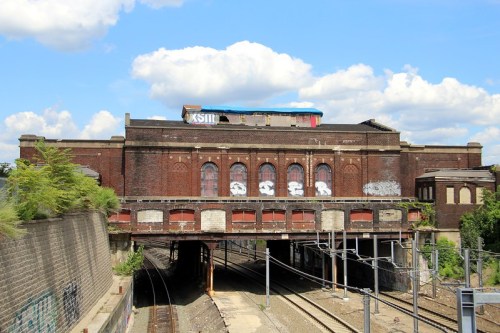 Abandoned brick building with arched windows, graffiti, and railway tracks below, set against a blue sky with clouds.