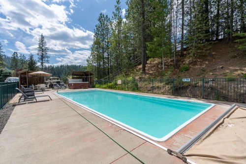 A clear swimming pool surrounded by trees and lounge chairs, with a hot tub nearby under a blue sky.
