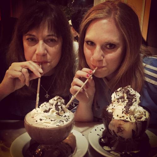 Two women sipping large ice cream desserts with whipped cream and chocolate, looking playfully at the camera.