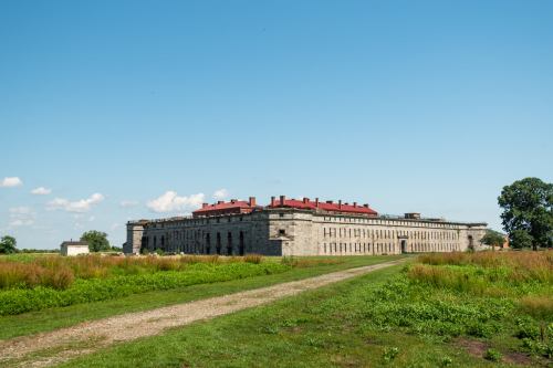 Historic stone building with a red roof, surrounded by green fields and a clear blue sky. Pathway leads to the structure.