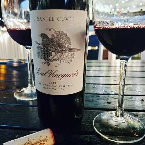 A bottle of J. Daniel Cuvée 2013 Cabernet Sauvignon from Lail Vineyards, with two glasses of red wine on a wooden table.