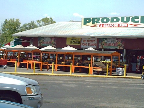 A colorful produce market with outdoor displays, umbrellas, and signs for fresh flowers and food stamps.