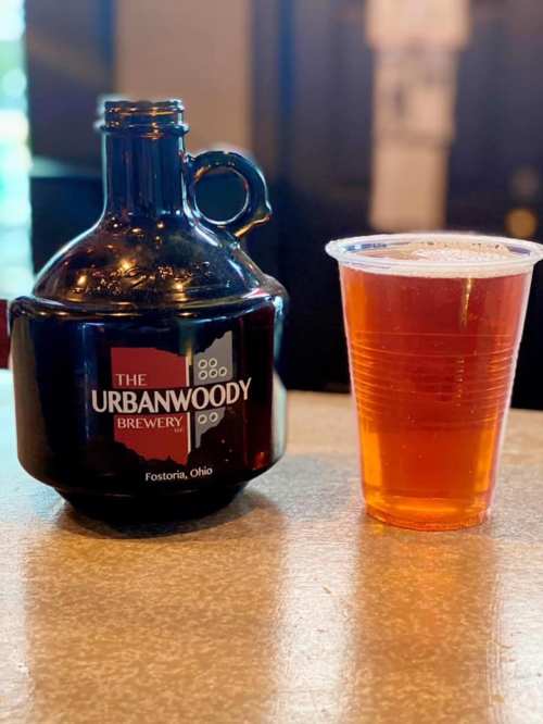 A brown growler labeled "The Urbanwoody Brewery" next to a clear cup of amber beer on a table.