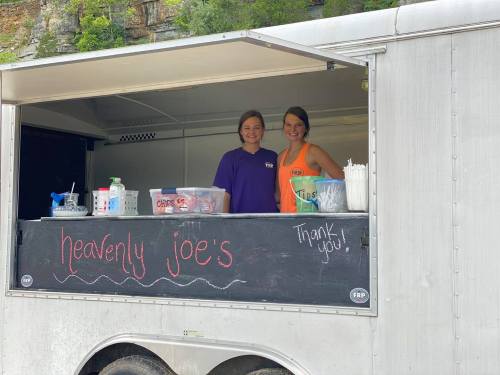 Two women smile from a food truck window labeled "Heavenly Joe's," with drinks and snacks visible inside.