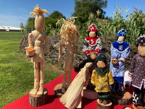 A display of handmade corn husk dolls in various colorful outfits, set against a green outdoor background.