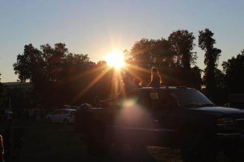 Silhouetted figures sit on a truck as the sun sets behind trees, creating a warm, glowing atmosphere.