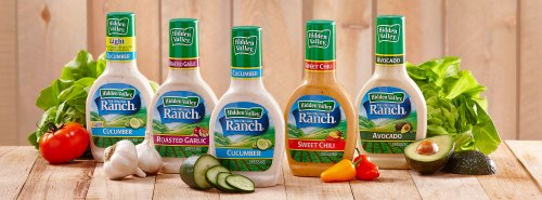 Five bottles of Hidden Valley Ranch dressing in various flavors, displayed on a wooden table with fresh vegetables.