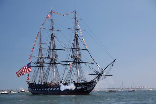 A historic tall ship sails on the water, adorned with flags and an American flag, with boats in the background.