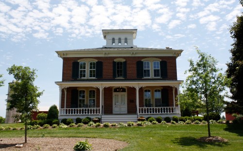A large brick house with a white porch, surrounded by green grass and trees, under a partly cloudy sky.