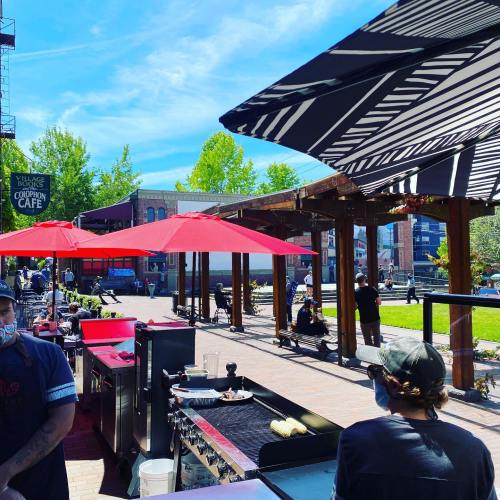 Outdoor café scene with red umbrellas, food prep stations, and people enjoying the sunny day in a vibrant setting.