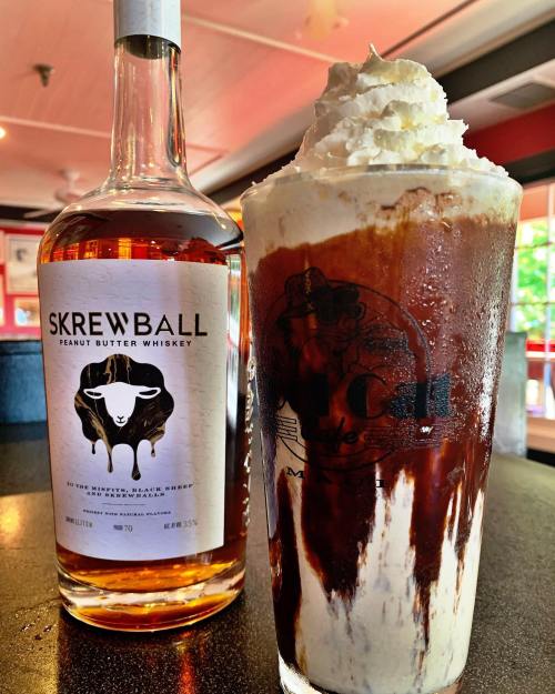 A glass of dessert topped with whipped cream and chocolate syrup, next to a bottle of Skrewball Peanut Butter Whiskey.